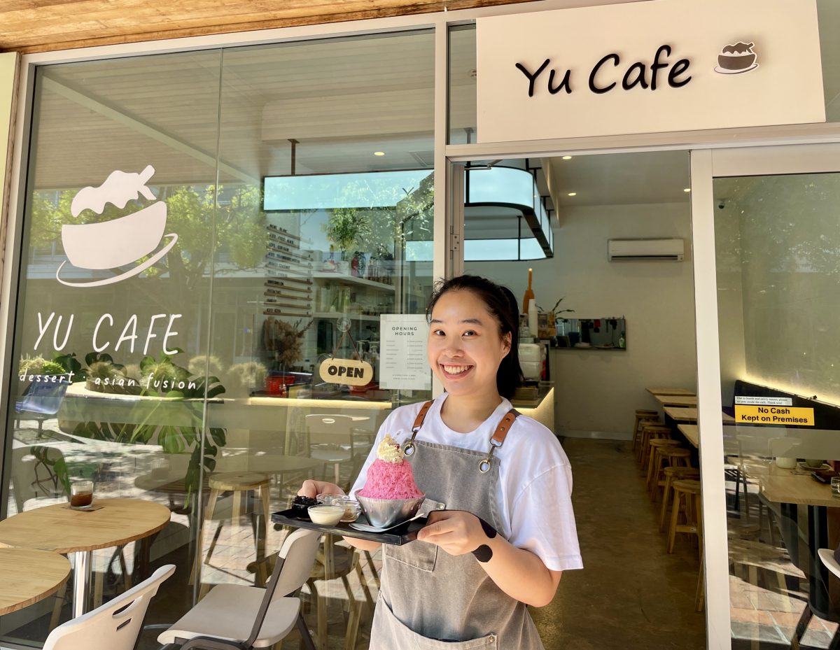 A young Asian woman in white t-shirt and grey apron stands in front of Yu Cafe. She holds a tray with a bright pink ice dessert. The dessert is topped with whipped cream, and the tray has ramekins of jelly and fruit. 