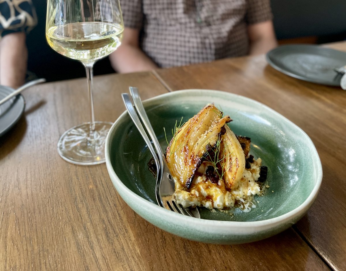 A shallow green bowl with wedges of caramelised fennel on creamy cheese. A glass of riesling is next to it.