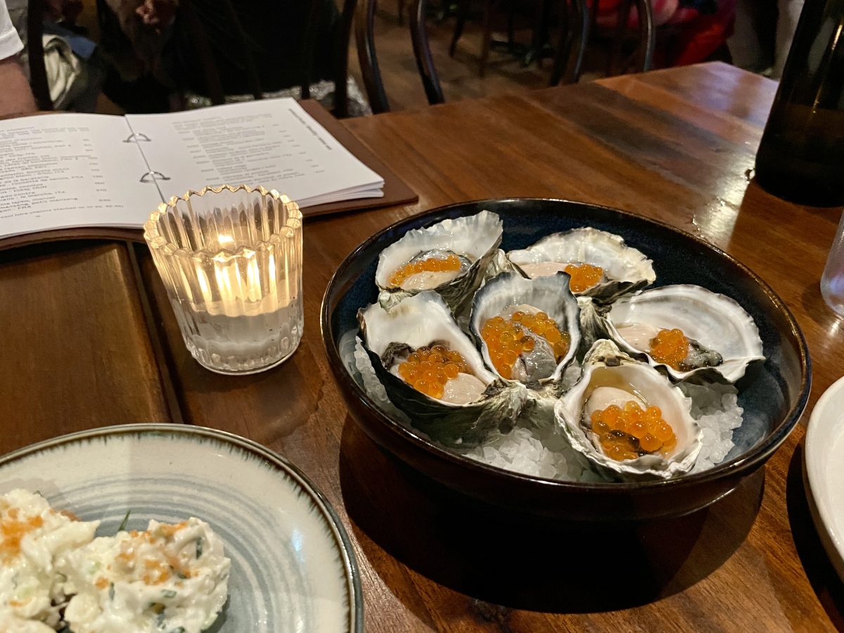 Six large oysters topped with orange salmon roe are served on rock salt in a black bowl.
