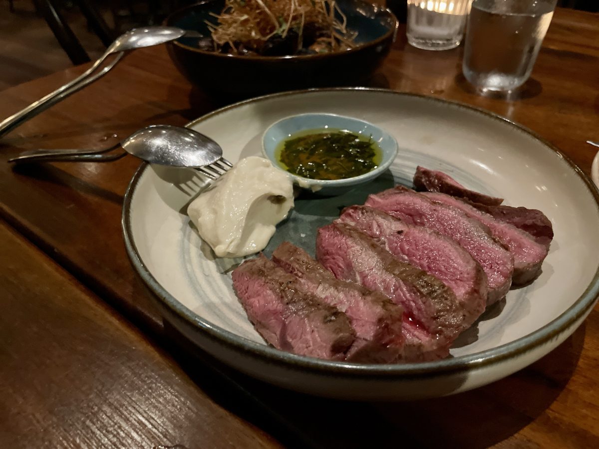 a plate with sliced, rare lamb, a dollop of a creamy looking dip and saucer of oil and herbs. In the background, a plate of mushrooms with crispy leek.