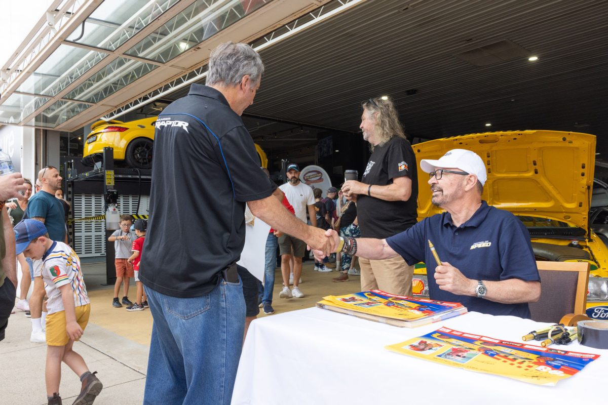 John Bowe sitting at a table signing posters, shaking hands with a person standing next to the table