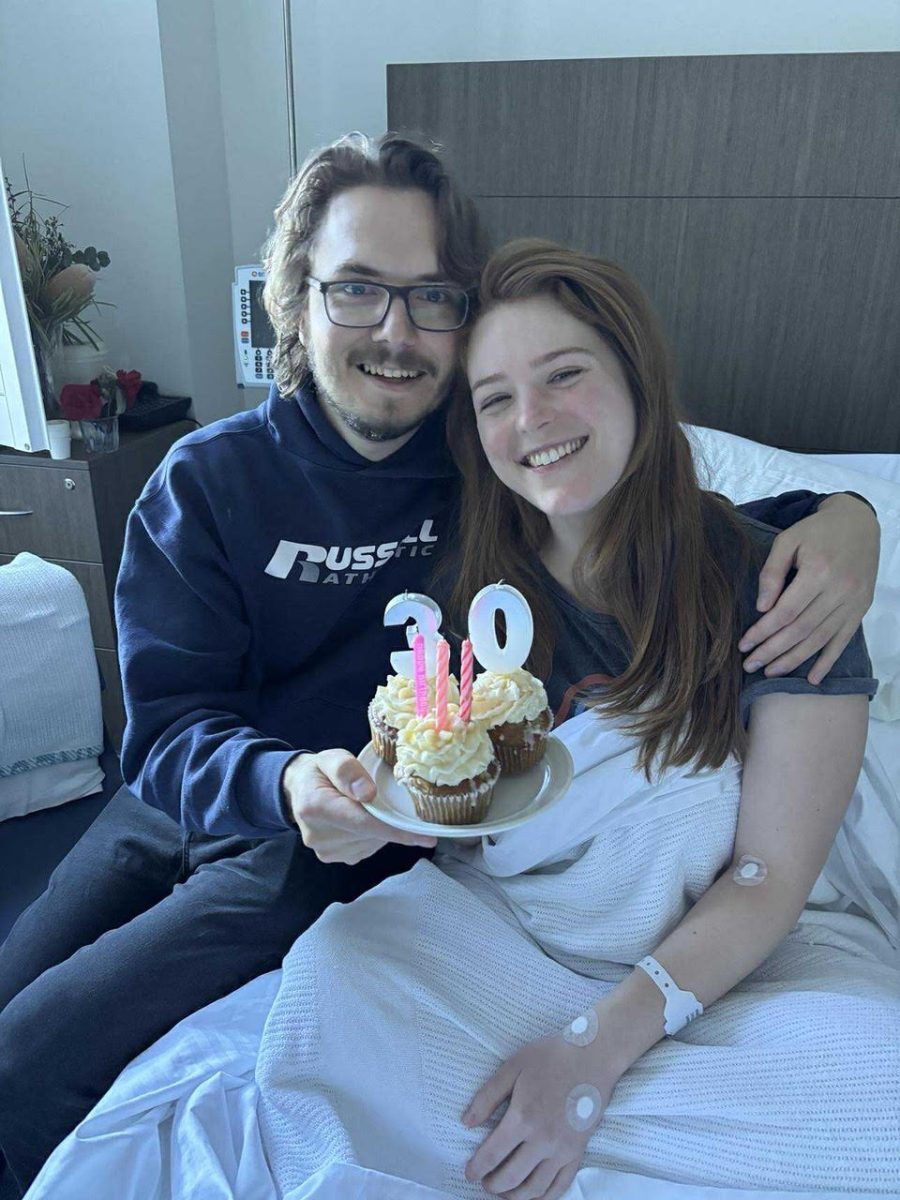 A young woman with red hair in a hospital bed. A young man has his arm around her and they are holding a plate of cupcakes with candles in the shape of the number 30.