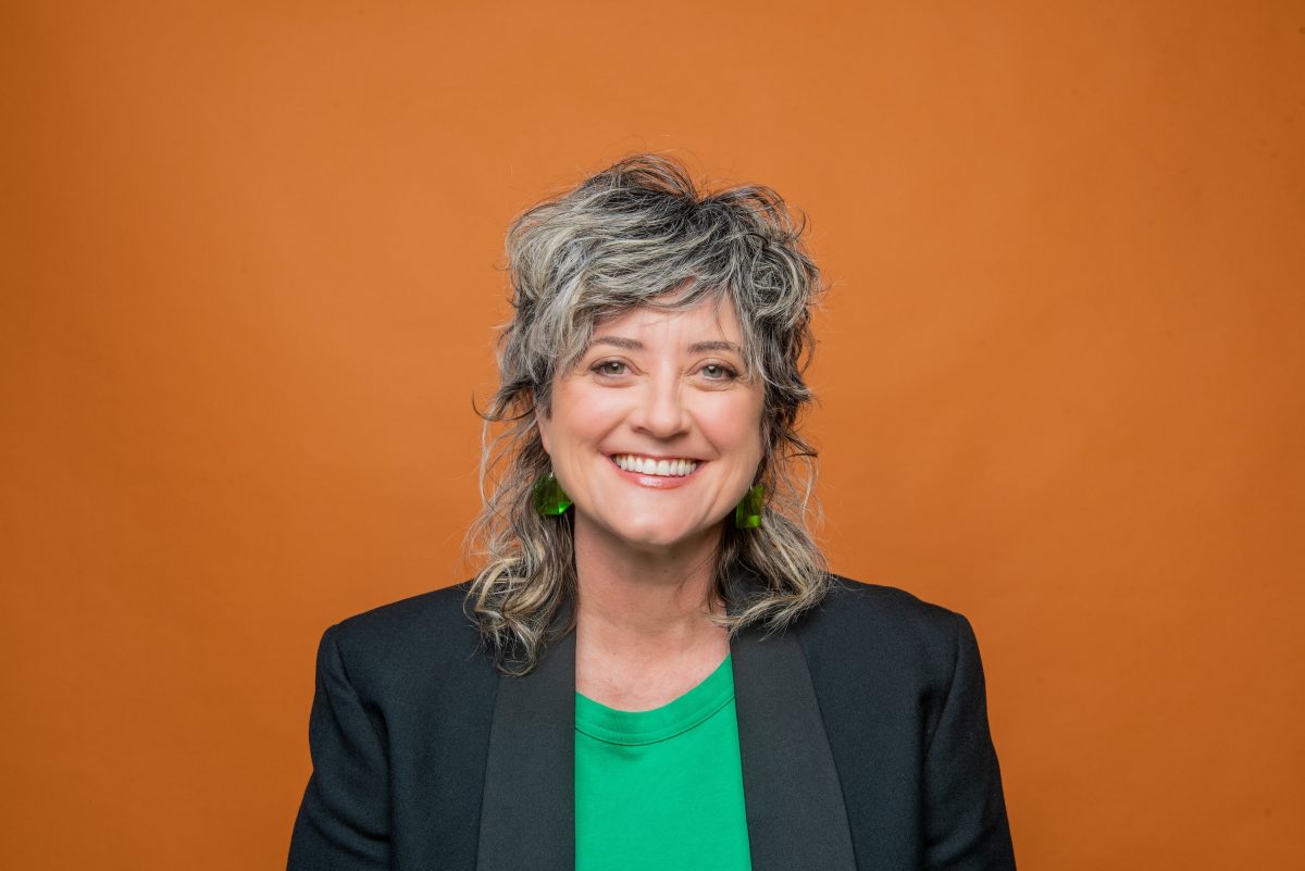 Comedian Chris Ryan stands in front of a bright orange background, smiling widely