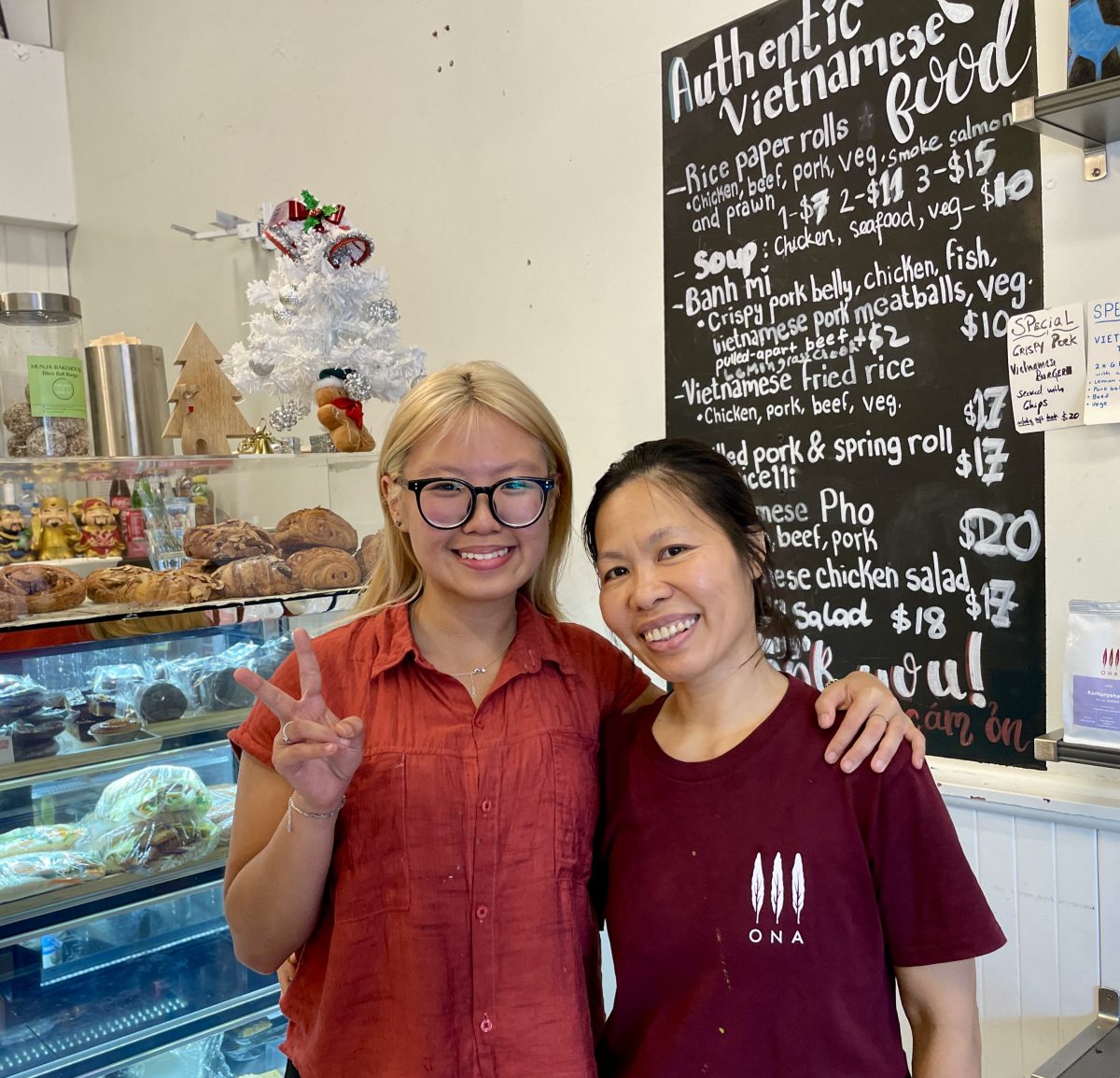 A young Asian woman with blonde hair and large glasses gives a peace sign with her fingers. She has her arm around an older Vietnamese women who is smiling and wearing an ONA branded shirt. Behind them is a blackboard menu reading 'Authentic Vietnamese Food' with menu items below.