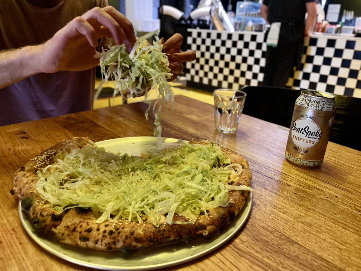 Hands picking up a piece of pizza covered in lettuce. Pieces of lettuce are falling off the piece.