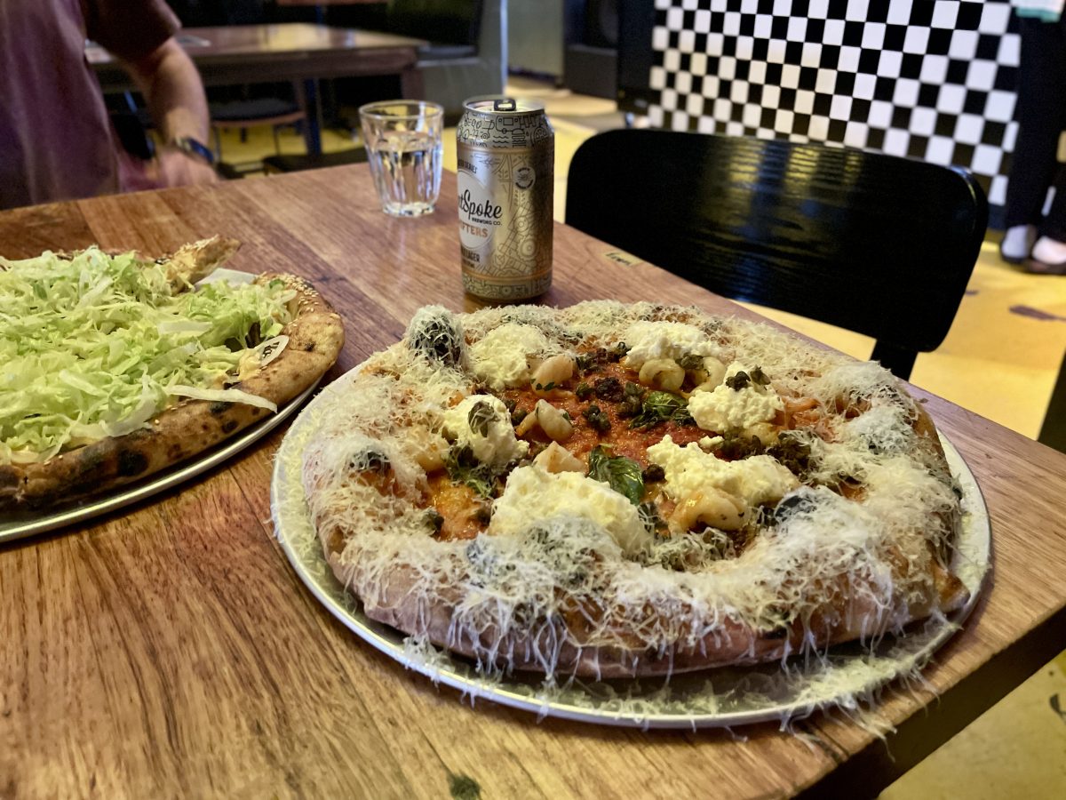 Two pizzas on a table with a can of beer behind. The pizza in the foreground has dollops of a creamy cheese, sprinkling of capers and prawns and lots of parmesan grated on top.