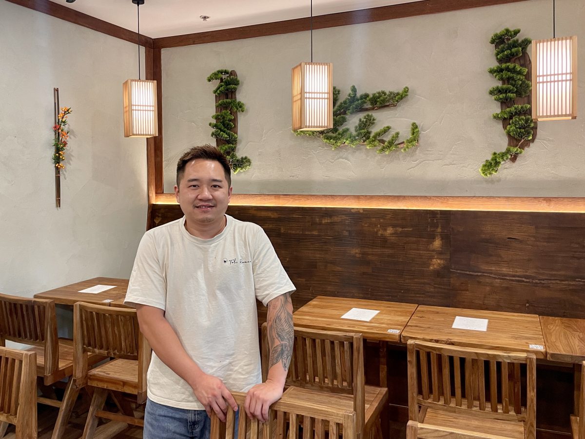 A man in white tshirt which reads 'Toli Ramen' stands in a dining room with wooden chairs and tables. 