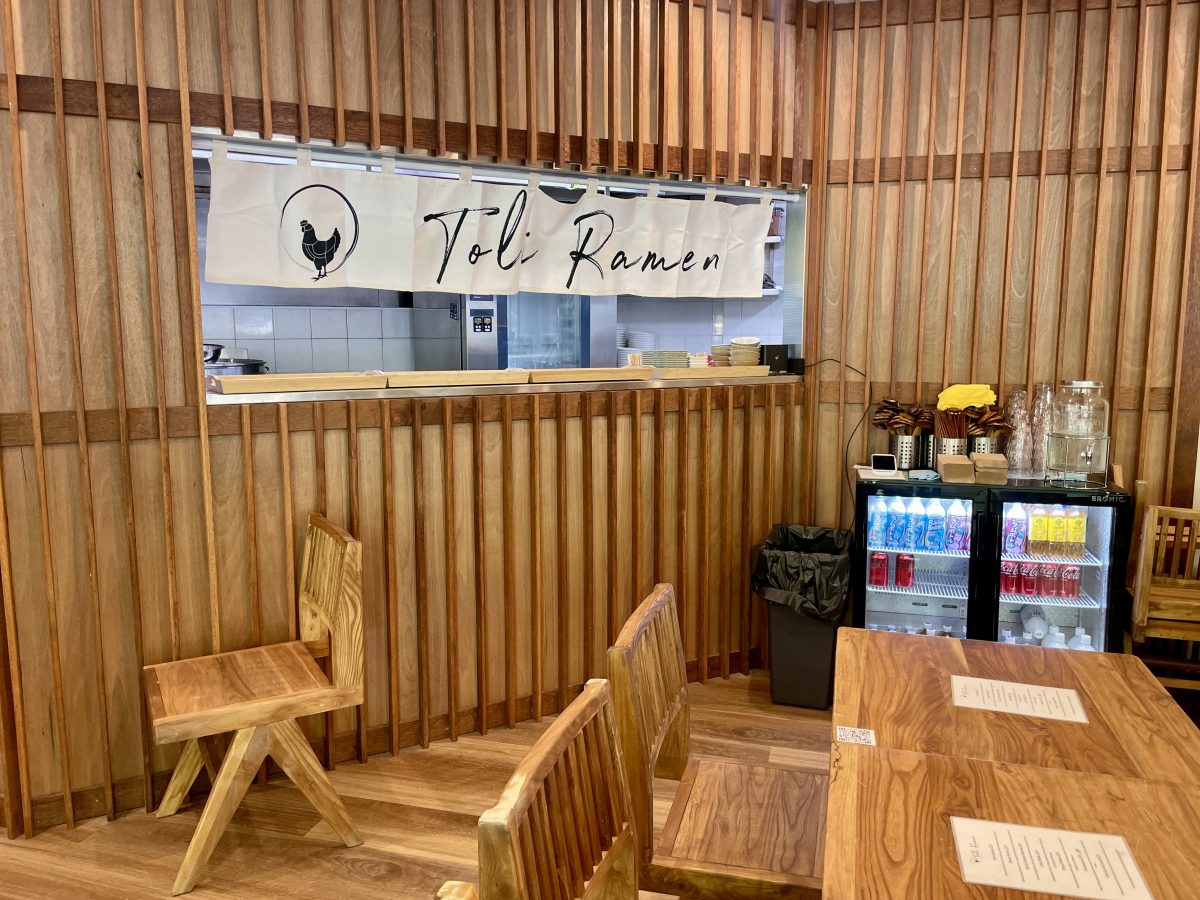 The dining room at Toli Ramen with view towards the kitchen pass, where there is a Toli Ramen banner, including logo of chicken in a circle.