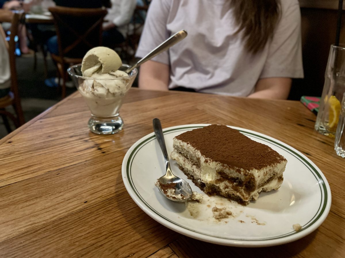 A large slice of tiramisu has one chunk taken out of the corner. In the background is a conical glass with scoops of icecream and a spoon.