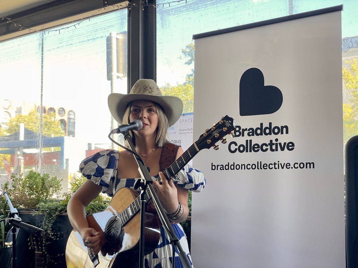 A young woman in blue and white dress and cowboy hat sings and plays guitar in front of a banner reading 'Braddon Collective'.