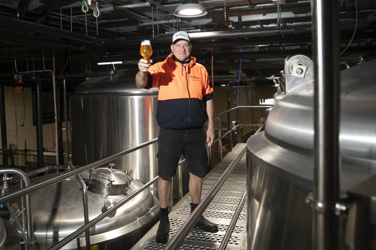 A man in a high vis shirt stands next to large stainless steel fermentation tanks. He is raising a glass of beer towards the camera.