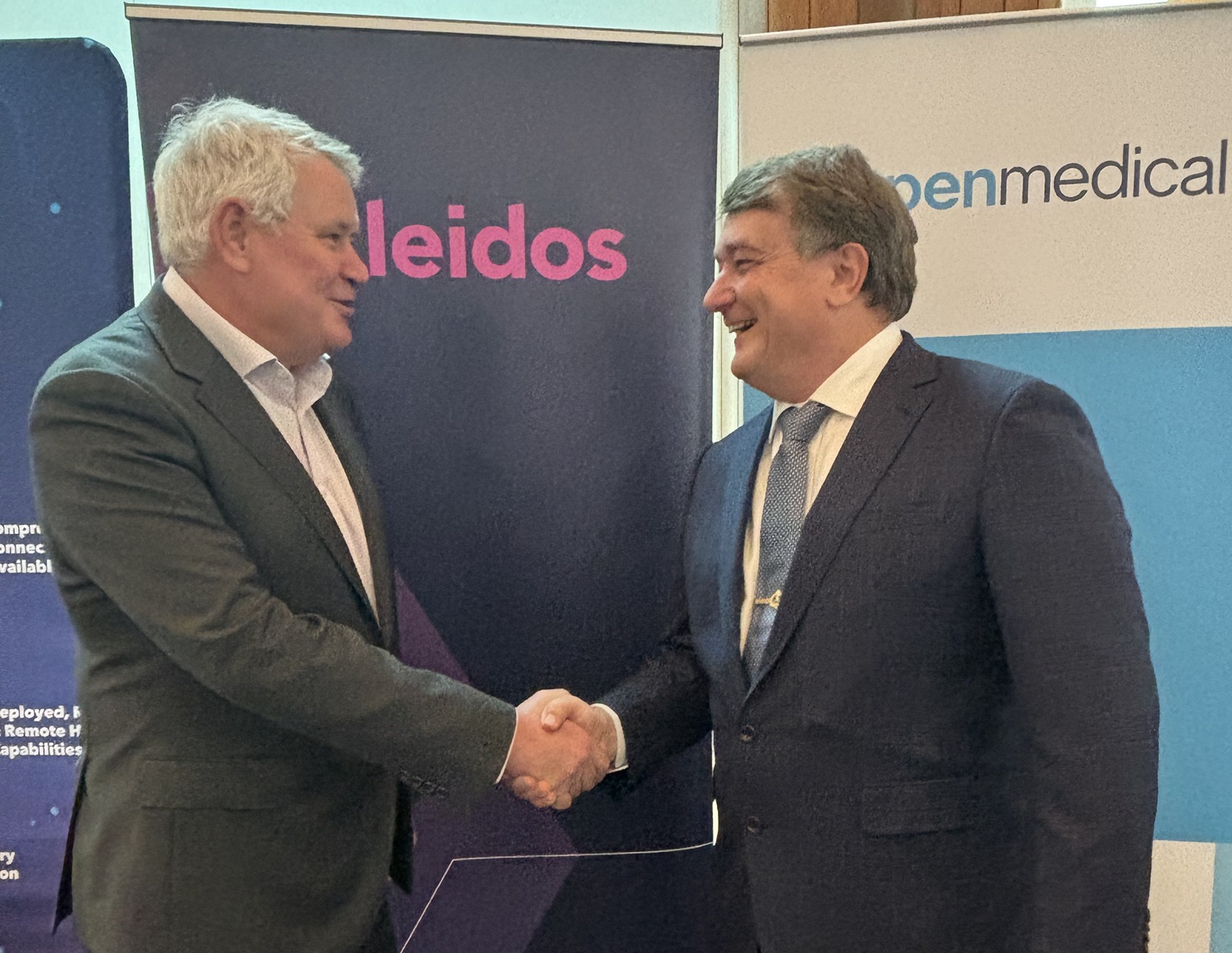 two business executive men shaking hands in front of company banners