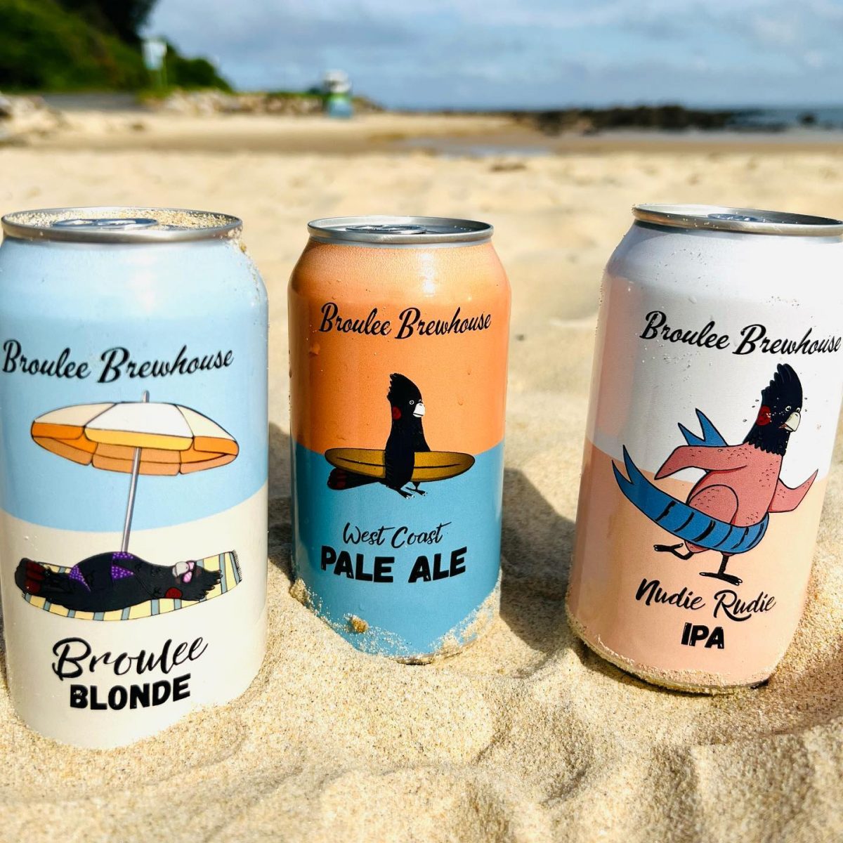 three cans of beer in the sand featuring a black cockatoo sunbathing, surfing and 'skinny dipping'