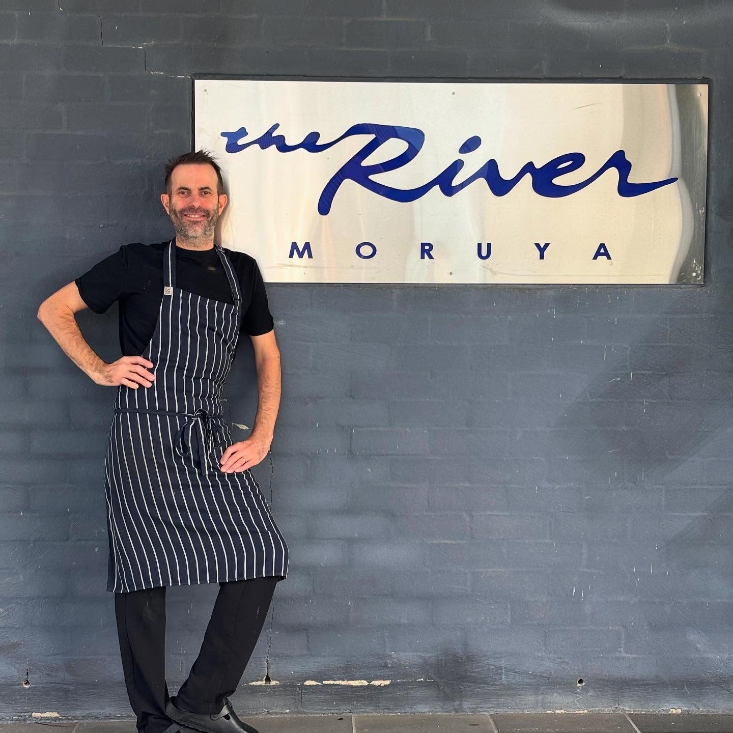 Peter Compton wearing black and a chef's apron stands next to a silver sign reading "The River Moruya" in blue flowing text.