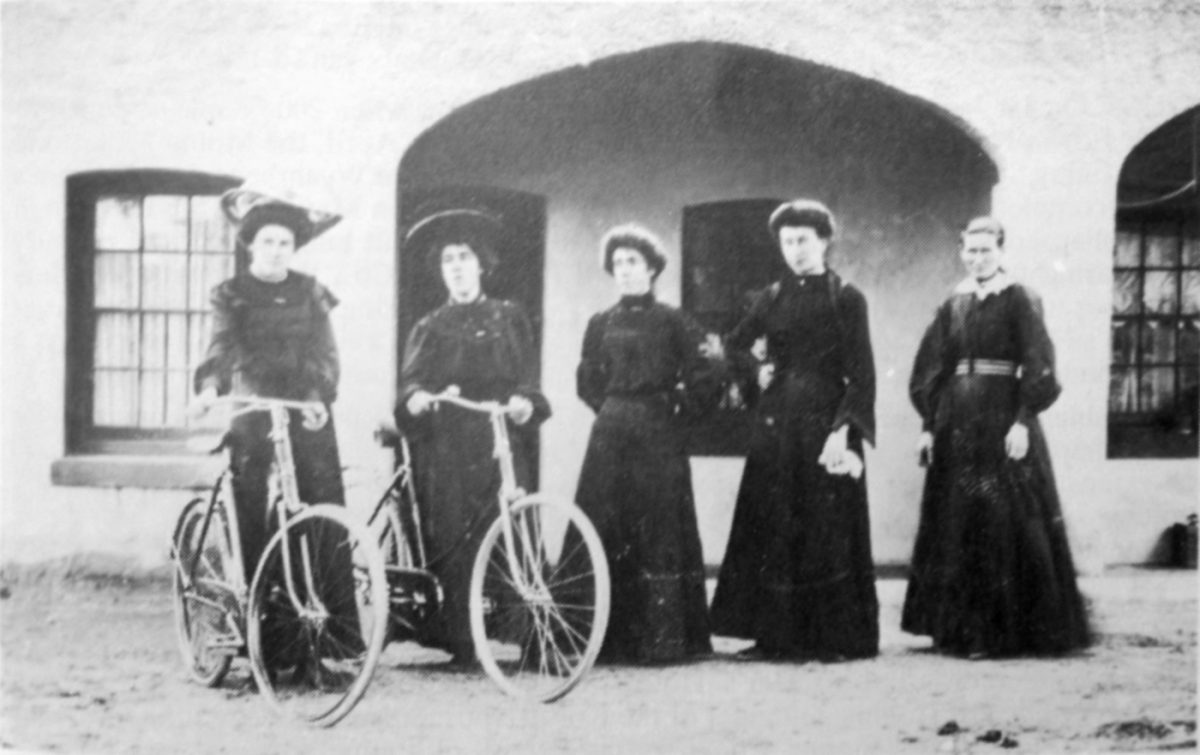 Victorian era women stand in front of building with arches.