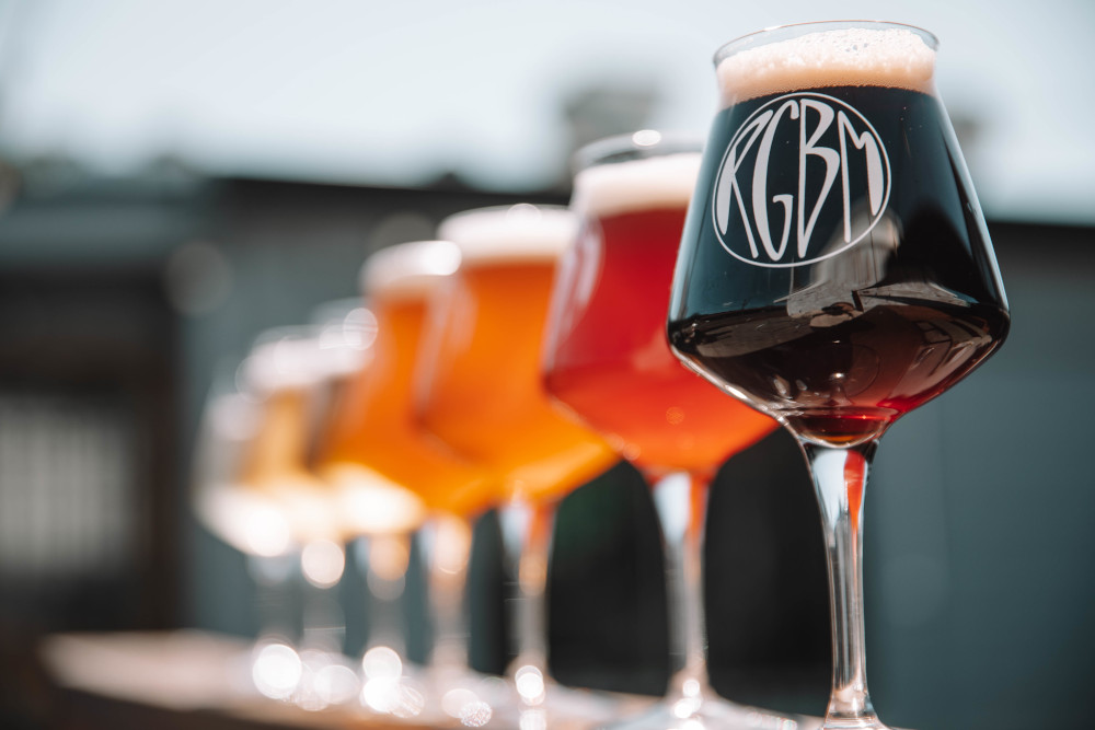 A shot of beers in stemmed glasses ranging from dark to light. A stylised logo with the letters RGBM is in focus on the nearest glass.